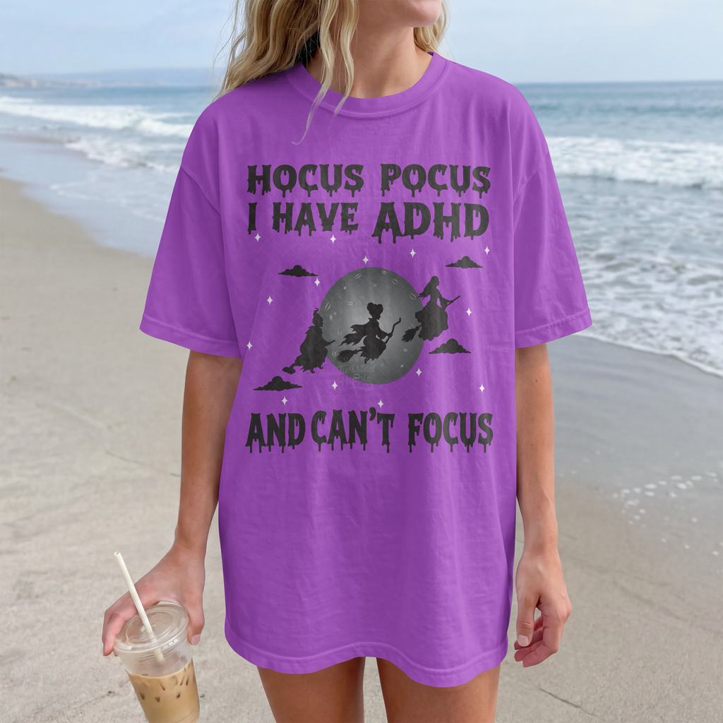 A woman standing on a beach wearing an oversized purple graphic T-shirt with a dark gray witch illustration and the text 'HOCUS POCUS I HAVE ADHD AND CAN'T FOCUS.' She is holding an iced coffee.
