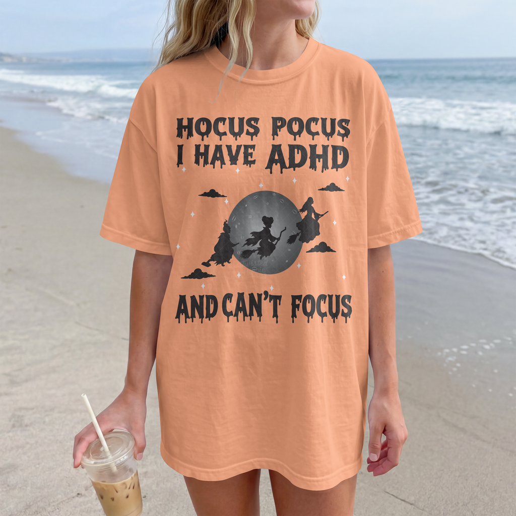 A woman standing on a beach wearing an oversized peach-colored graphic T-shirt with a dark gray witch illustration and the text 'HOCUS POCUS I HAVE ADHD AND CAN'T FOCUS.' She is holding an iced coffee.